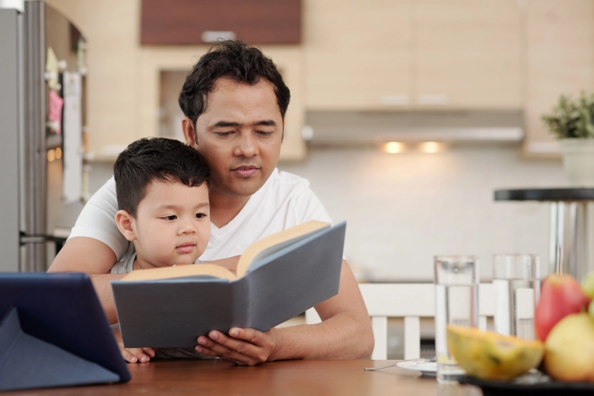Seorang ayah sedang membacakan buku kepada anak laki-lakinya, yang sudah terlindungi asuransi pendidikan sejak dini